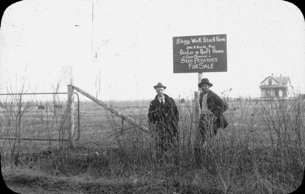 A handwritten sign identifies the "Shing-Wak Stock Farm, John B. Breselle (?) Prop., Dealer in Draft Horses." It also advertises "Green Mountain Seed Potatoes For Sale." An unidentified white man, left, is standing beside a Native American man who is presumably the farm's owner. In the background there are two animals grazing in the background, and on the right is a story and one half farmhouse with an open front porch.
