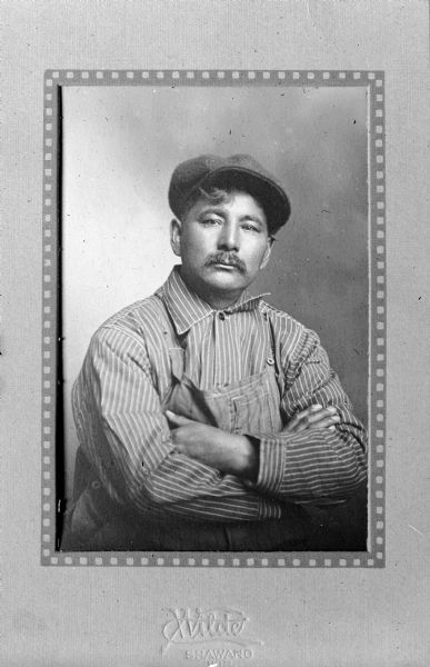 Waist-up seated studio portrait of Ernest Oshkosh (?1875-1929), grandson of Chief Oshkosh, son of Neopit. He is wearing a cap, bib overalls, and a striped work shirt. Ernest Oshkosh was a Menominee chief and agency farmer at Keshena from 1912 until his death in 1929. He worked closely with University of Wisconsin Extension administrator Joseph Wojta to improve agricultural practices of Native Americans in Wisconsin through education.