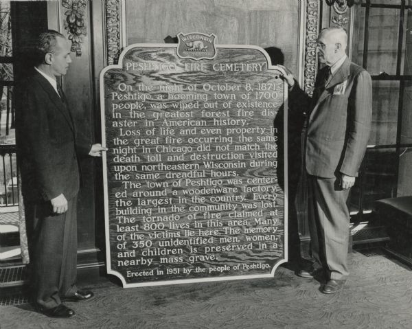 Governor Walter J. Kohler Jr., (left) and James R. Law, state highway commission chairman, standing indoors with the Peshtigo cemetery marker. The marker was dedicated Sunday, October 7, 1951, the 80th anniversary of the fire. 