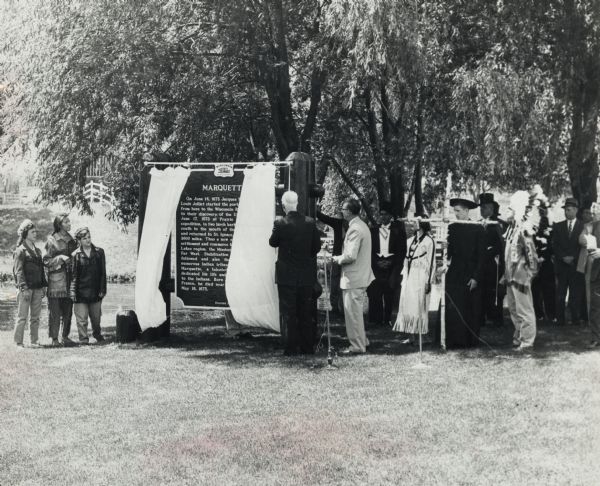 View across lawn towards a crowd, with costumed participants dressed as Native Americans, fur traders, and Jacques Marquette, at the dedication of the Marquette historical marker. Dedication festivities included a barbecue lunch, unveiling ceremony along the highway, parade, and pageant. 