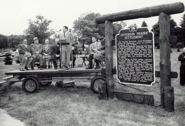 Judge Chester R. Christensen, Rock County Judge, speaking at the dedication ceremony of the Jefferson Prairie Settlement historical marker. Seated behind him are other speakers at the ceremony. From left to right: Reverend E.B. Burtness, Capron Lutheran church, Reverend Marcus Lewis, Luther Valley church, Einar Hammer, president of Region 5 of Sons of Norway, Reverend O.M. Skindrud, Jefferson Prairie church, Reverend Olaf Lysnes (obscured by Judge Christensen speaking), Raymond S. Sivesind, supervisor of historic sites and markers, and Professor Robert Richardson of Beloit College. The group of men are sitting and standing on a trailer parked near the marker.