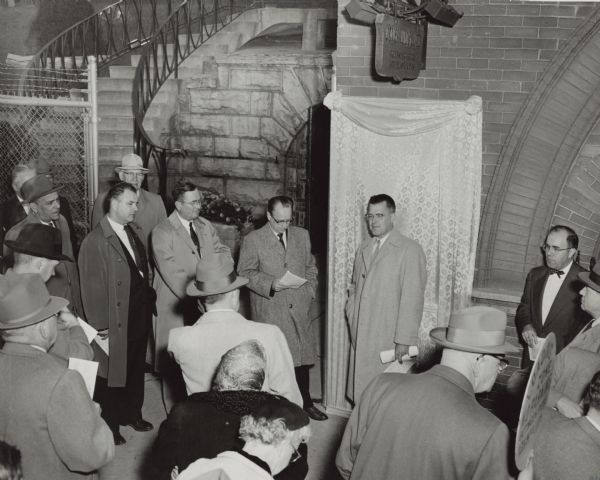 Raymond Sivesind, director of the sites and markers program of the State Historical Society, as a speaker in the dedication and unveiling ceremony, stands in front of the covered Watertown Plank Road historical marker. Other speakers include, on the right, Frederick I. Olson, president of the Milwaukee Historical Society, Norman R. Klug, to the left of Sivesind, president of the Miller Brewing Company, and next to Klug, James Fitzpatrick, president of the Watertown common council. There is a sign for the Miller Brewing Co. Caves above the marker. 