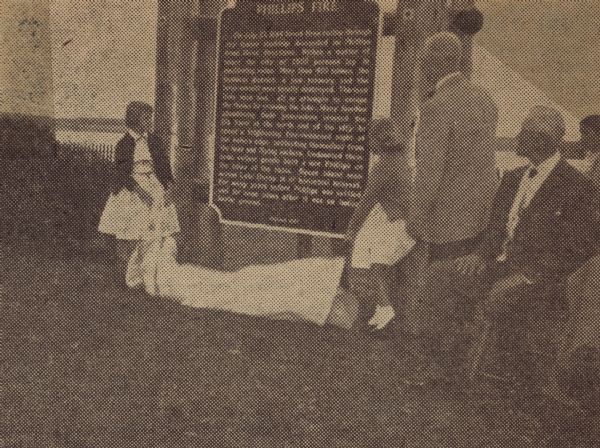 Mary Lynn Norton and Cathy Mess unveiling the Phillips Fire historical marker, while Avrid Bostrom and Alderman Victor Cress (seated) look on. Bostrom, a local jeweler, headed the Lakeside Park planning and development, and was instrumental in acquiring the historical marker for the park. Cress was a survivor of the terrible fire and spoke about the day the fire raged in 1894.  