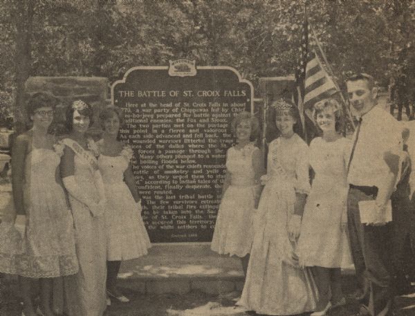 The newly crowned beauty queens and attendants from St. Croix Falls and neighboring Taylors Falls stand with Willis Erickson (right), president of the Polk County Historical Society and master of ceremonies, at the dedication of The Battle of St. Croix Falls historical marker. Ruth Ann Peterson, Queen Margaret Thomas, and Barbara Larson, from St. Croix Falls, are standing on the left, while the girls from Taylors Falls, Julie Olson, Queen Christine Johnson, and Julie Vitalis are standing on the right next to Erickson. The girls looked on as local Boy Scouts unveiled the marker to the public. 