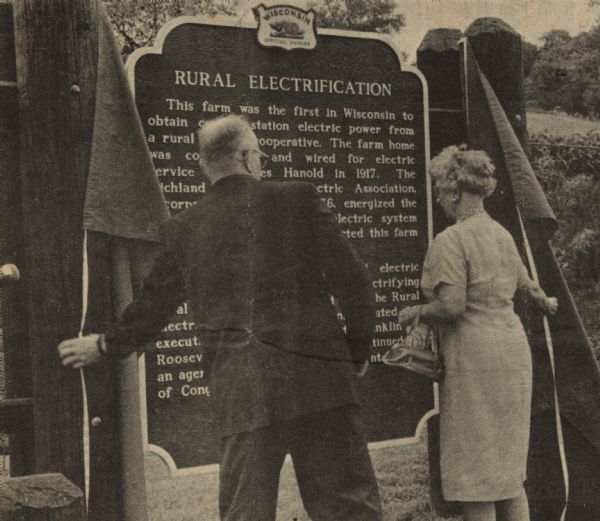 Mrs. A.V. Miller and James R. Sullivan unveiling the Rural Electrification historical marker together at the dedication ceremony outside of Richland Center. Mrs. Miller's late husband served as the first president of the Richland Cooperative Electric  Association, and Mr. Sullivan, the assistant administrator of the Rural Electrification Administration, made the trip from Washington D.C. to be at the dedication of the historical marker. 