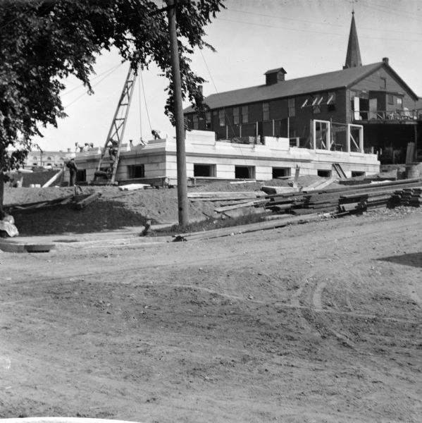 View across unpaved road towards construction workers working on the first floor of the Eau Claire Public Library at the intersection of Farwell Street and Grand Avenue. The Carnegie library was designed by the firm Patton & Miller of Chicago and opened in 1904. In the background behind a building on the right is what may be a church steeple.