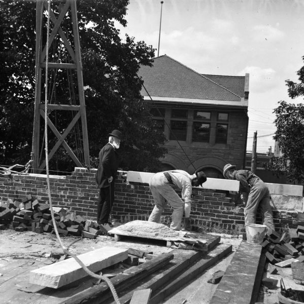 Two men are laying bricks during the construction of the Eau Claire Public Library at Farwell Street and Grand Avenue. A third man, at left, wearing a hat and suit, and holding a cane, stands nearby watching their work. In the foreground is a rope which is attached to a wooden structure supporting a winch and pulley set up behind the brick wall on the left. The library, designed by the firm Patton & Miller of Chicago, opened in 1904.