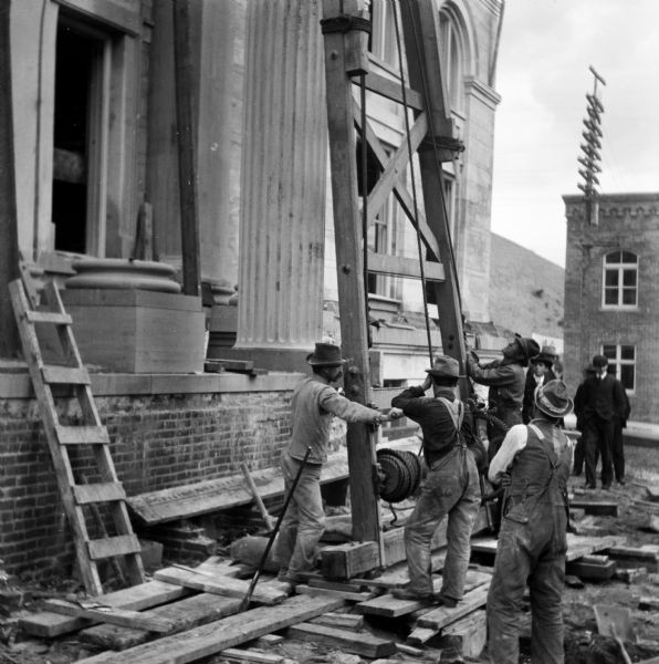 View of a group of construction workers standing on the side of the building on a stack of lumber. They are using a large wooden structure supporting a pulley and winch during the installation of columns at the Eau Claire Public Library at Farwell Street and Grand Avenue. The library opened in 1904. A group of men wearing suits and hats are standing and watching in the background.