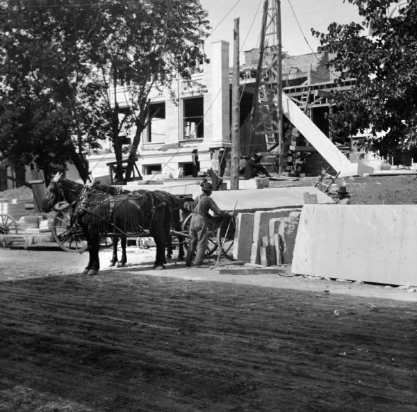 View across unpaved road toward construction workers loading a stone slab onto a horse-drawn wagon during the construction of the Eau Claire Public Library at Farwell Street and Grand Avenue. In the background, men are standing near a large wooden winch and pulley set up at the partially constructed library.