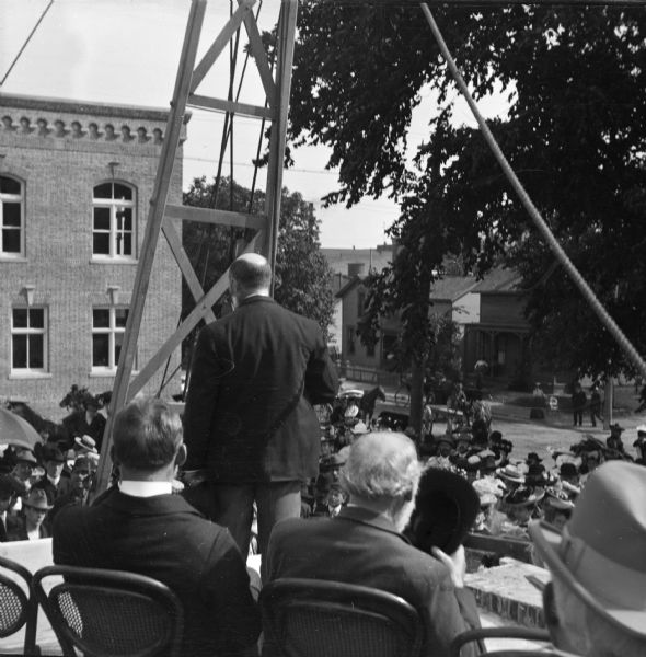 View from back of stage toward men sitting in chairs listening to a man standing and speaking at the ceremony at which the cornerstone for the Eau Claire Public Library was laid during construction. There is a crowd below the stage, and more people are standing on the street and sidewalk in the background. The Carnegie library opened the following year. There is a large wooden structure supporting a winch and pulley system set up near the front of the stage.