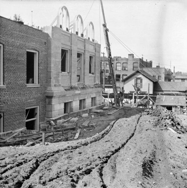 View from the rear of the Eau Claire Public Library during its construction. A construction worker stands at the base of a wooden support structure with a winch and pulley. Frames for arched windows are being placed in the upper floor of the library at Farwell Street and Grand Avenue. In the background is a horse-drawn vehicle on the street below, and buildings can be seen in the far background.