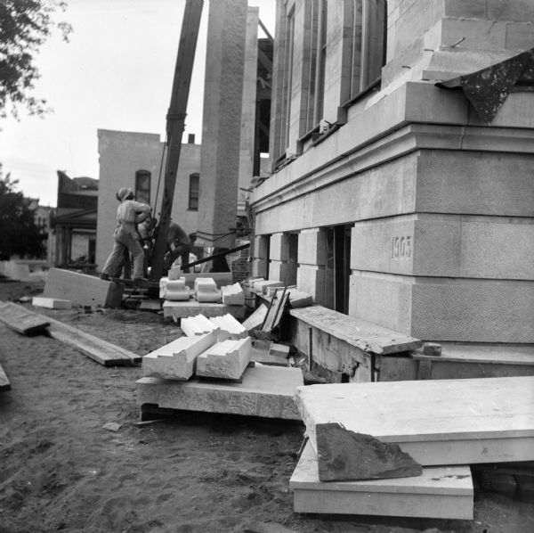 View from corner of new library building toward a group of construction workers lifting stonework into place with a large wooden structure supporting a winch and pulley system during the construction of the Eau Claire Public Library at Farwell Street and Grand Avenue. In the foreground the cornerstone reads "1903."