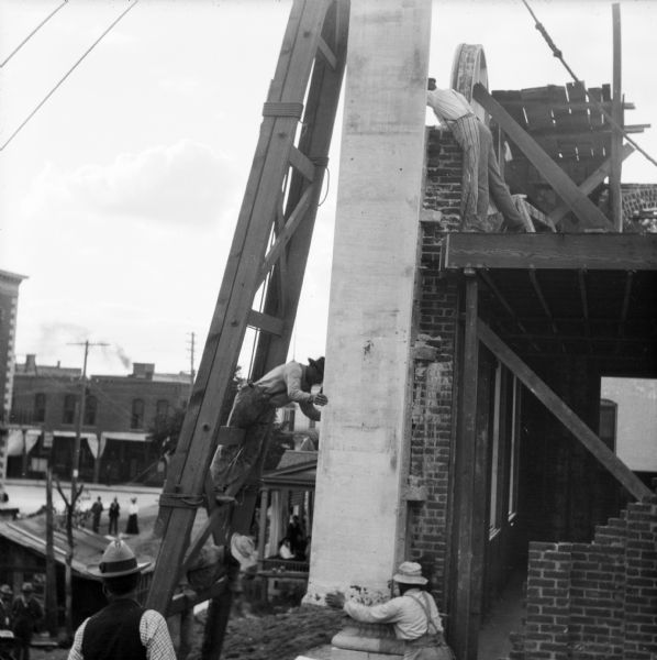 View of group of construction workers lifting stonework into place with a large wooden structure supporting a winch and pulley system during the during the construction of the Eau Claire Public Library at Farwell Street and Grand Avenue. In the background below people are watching from the porch of a house and from the street.