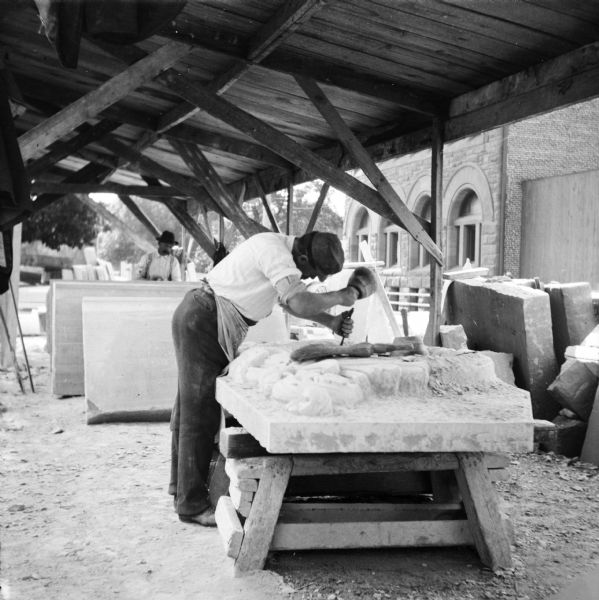 A construction worker is chiseling a decorative element on a stone slab during the construction of the Eau Claire Public Library at Farwell Street and Grand Avenue. Another man is standing in the background with slabs of stone. Both men are working underneath a roofed, open-sided wood structure.