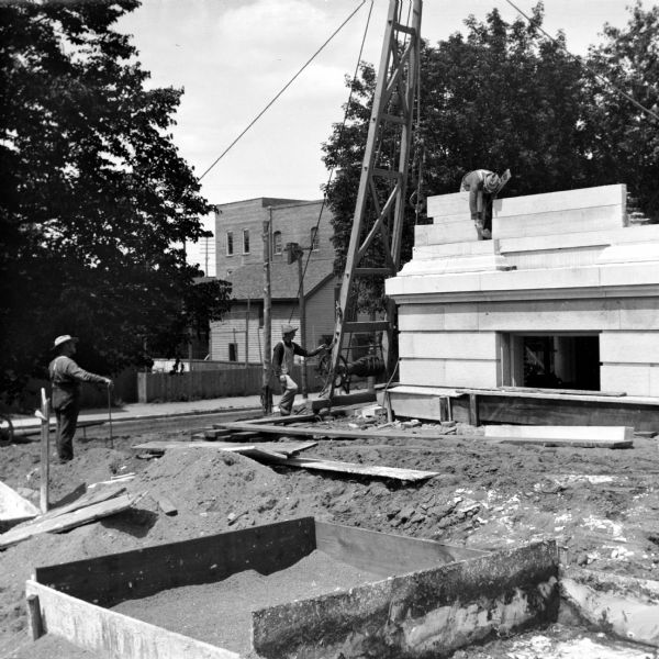 Construction workers are laying stone on the facade of the Eau Claire Public Library at Farwell Street and Grand Avenue. One man is standing on the first floor, another man is standing at the base of a wooden structure supporting a winch and pulley set up at the corner of the building. A third man stands watching on the left among piles of dirt and lumber. There are buildings across the street in the background.