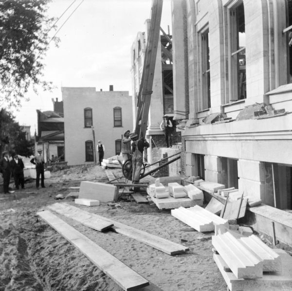 Construction workers are lifting a piece into place using a wooden structure supporting a winch and pulley set up near the facade of the Eau Claire Public Library, while men are watching from the grounds and the building. 