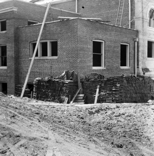 View across dirt grounds towards the Eau Claire Public Library at Farwell Street and Grand Avenue. Roof tiles are stacked at the corner of the brick section of the building during its construction. A section of the stone facade is on the far right.