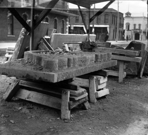 Stone slabs under a roofed, open-sided wood structure at the construction site of the Eau Claire Public Library at Farwell Street and Grand Avenue. The slab is in the process of being carved. Other slabs of stone are leaning against one of the beams. In the background are buildings across the street.