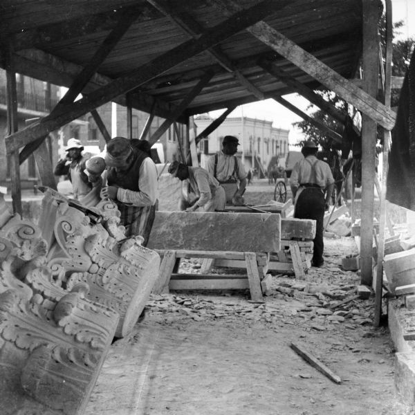 A worker under a roofed, open-sided wood structure is chiseling details in the capital of a Corinthian column, at left, while behind him other men are working on large stone pieces during the construction of the Eau Claire Public Library at Farwell Street and Grand Avenue. In the background, horse-drawn vehicles and pedestrians are in the street near commercial buildings. 