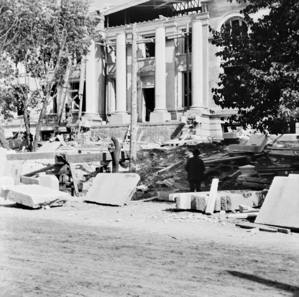 View from street toward the partially completed construction of the Eau Claire Public Library at Farwell Street and Grand Avenue. A man is standing and watching on the right, and men are working in front of the building to the left of the entrance with a wooden structure supporting a winch and pulley. The four columns have been put in place, but they do not yet have their capitals.