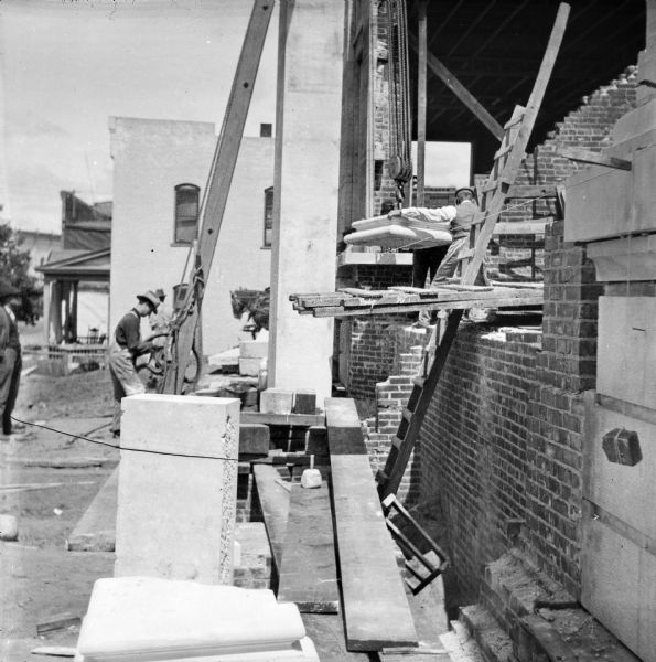 View along side of building toward construction workers moving materials during the construction of the Eau Claire Public Library at Farwell Street and Grand Avenue. On the far left a man is watching, and one man is working at the base of a wooden structure supporting a winch and pulley. Another man is standing on the first floor and helping to position what may be the stone base for a column.