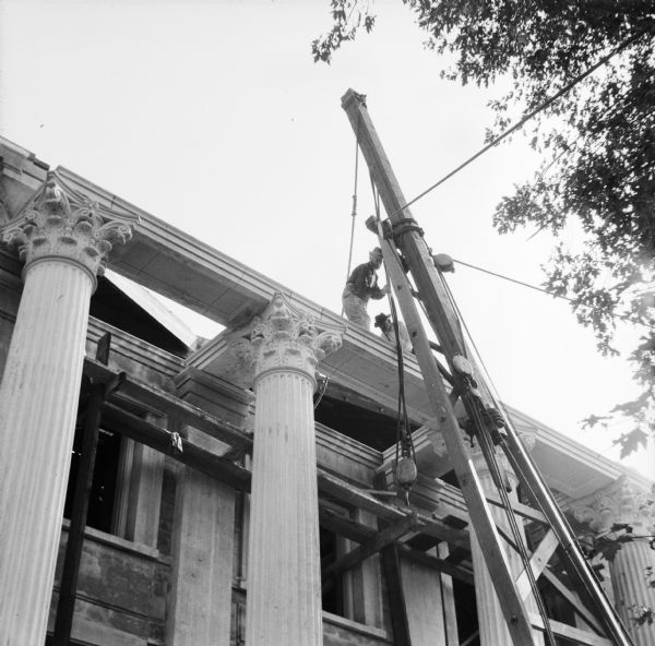 View looking up toward construction workers on the roof above the columns of the Eau Claire Public Library during its construction. The men are guiding a rope and pulley which is attached to a tall, wooden structure supporting a block and tackle system.