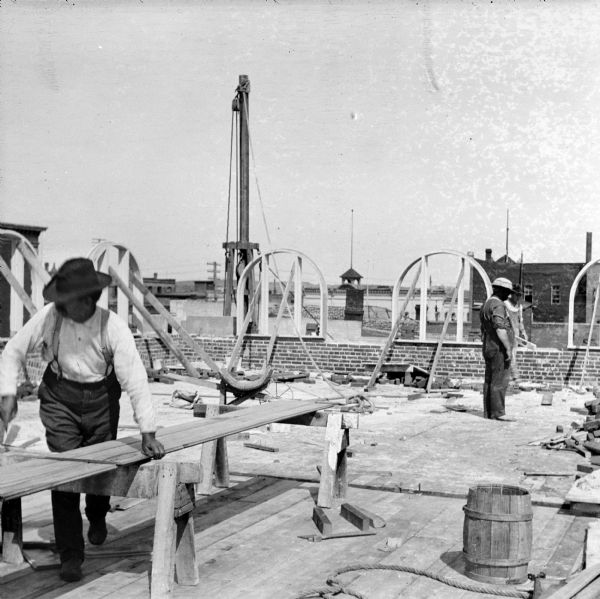 Two workers on one of the upper floors of the Eau Claire Public Library during its construction. The frames of the arched windows are in the background. The block and tackle system is behind the brick wall in the background.