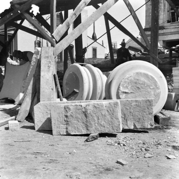 Portions of columns awaiting installation are stacked near a wooden open-sided building during the construction of the Eau Claire Public Library at Farwell Street and Grand Avenue. A man is working on one of the stone capitals, and another man is sitting in the shade in the center. In the background on the left is the stone facade of the partially constructed library building.