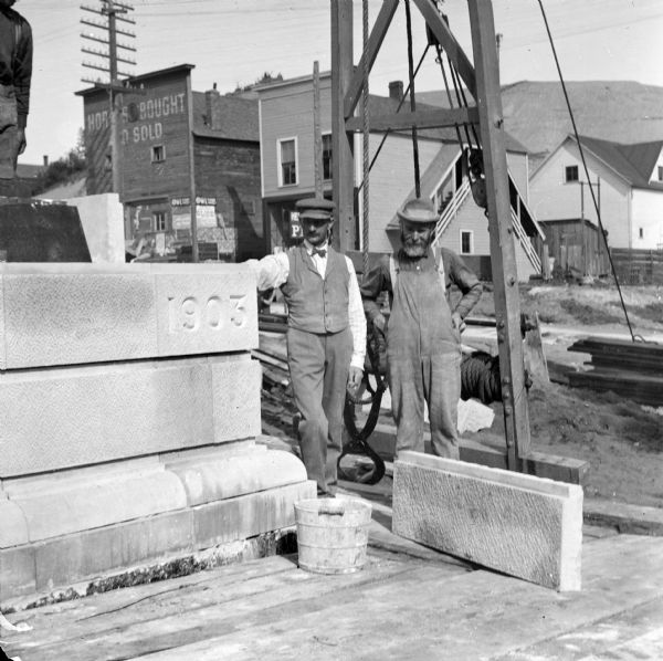 Two men are standing and posing beside the cornerstone of the Eau Claire Public Library. A third man is standing on the far left just above and behind the cornerstone, which was laid on June 16, 1903. The Carnegie library, at Farwell Street and Grand Avenue, opened in 1904. Construction equipment, including a winch and pulley on a wood structure is directly behind the two men, and commercial buildings are across the street in the background. There is a large hill in the far background behind the buildings.