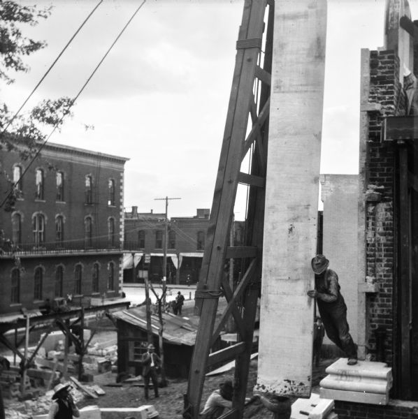 View looking down toward construction workers moving a stone slab for the facade into place during the construction of the Eau Claire Public Library at Farwell Street and Grand Avenue. The workers are using a using a tall, wooden structure supporting a winch and pulley, or block and tackle. People are watching from below. Across the street on the left are commercial buildings.