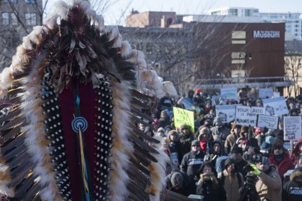 Save the Mounds demonstration around the Capitol Square against Assembly Bill 620. In the foreground, seen from behind, is the headdress worn by Chief Clayton Winneshiek of the Ho Chunk Nation. Below in the background is a crowd, with many holding protest signs. In the far background is the Wisconsin Historical Museum building at the top of State Street.