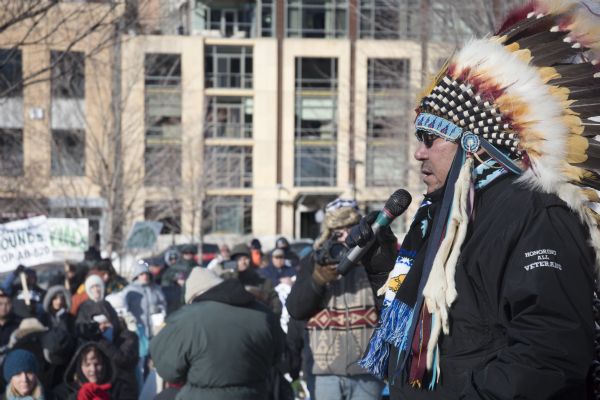 Save the Mounds demonstration around the Capitol Square against Assembly Bill 620. Chief Clayton Winneshiek of the Ho Chunk Nation, wearing a headdress, is speaking into a microphone. In front of him is a crowd, with some protestors holding signs.