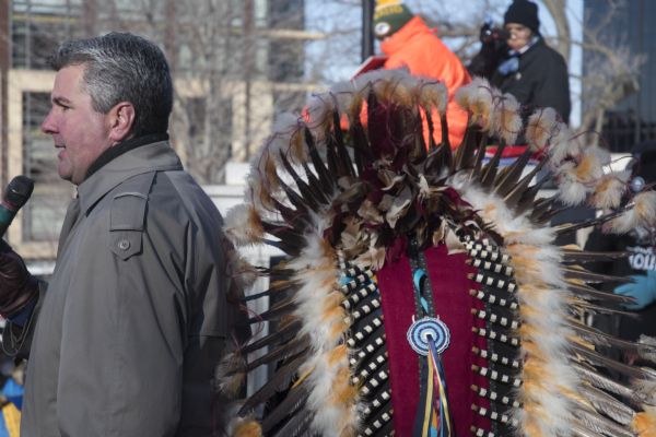 Save the Mounds demonstration at the Capitol Square against Assembly Bill 620. Wisconsin State Representative Robb Kahl is standing on the left speaking into a microphone. Behind him is a headdress, seen from the back, worn by Chief Clayton Winneshiek of the Ho Chunk Nation, who introduced Mr. Kahl.