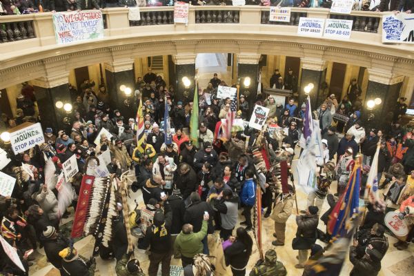 View looking down at the Save the Mounds demonstration on the ground floor of the Wisconsin State Capitol against Assembly Bill 620. In the center is a drum circle. Surrounding the drummers are demonstrators holding, flag, standards and signs. Above, signs are attached to the second floor railing.