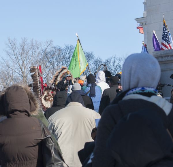 Save the Mounds demonstration around the Capitol Square against Assembly Bill 620. Chief Clayton Winneshiek of the Ho Chunk Nation, wearing a headdress, is speaking into a microphone. Surrounding him is a crowd, with some protestors holding standards and flags.