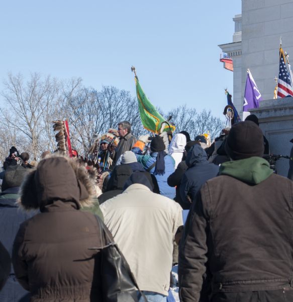 Save the Mounds demonstration at the Capitol Square against Assembly Bill 620. Wisconsin State Representative Robb Kahl is standing in the center speaking into a microphone. Behind him is Chief Clayton Winneshiek of the Ho Chunk Nation, wearing a headdress. They are surrounded by a crowd of protestors holding standards and flags.