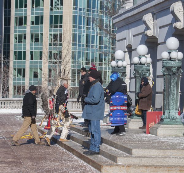 Save the Mounds demonstration around the Capitol Square against Assembly Bill 620. A Native American man dressed in traditional clothing walks up the steps towards the Capitol door. A policeman looks on. A woman wears a blanket with a traditional motif. In the background are office buildings.
