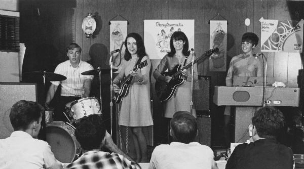 View from audience of the band "The Paraphernalia" performing. From the left is: a young man is playing the drums, two young women each playing an electric guitar, and a woman playing keyboards.