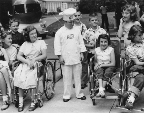 Meinhardt Raabe as Little Oscar standing with a group of children, some in wheelchairs, at Easter Seals Camp Wawbeek. The Wienermobile is in the background on the left.