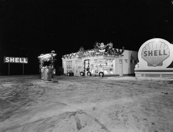 Night view of a Shell service station with Christmas decorations. There are tree branch swags on the building and on the poles and light fixture around the pumps. On the roof above the decorated windows of the station is a cut-out of Santa Claus and pine trees.