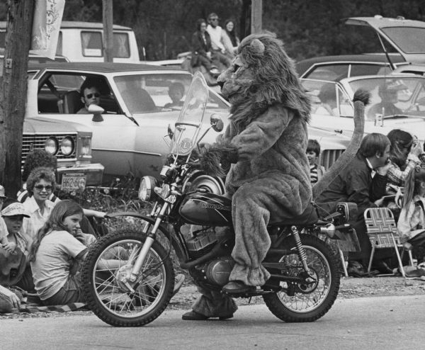 A person costumed in a Lion suit is riding a motorcycle as part of the Edgerton Lions Club. Onlookers are sitting along the side of the road and in parked cars in the background.