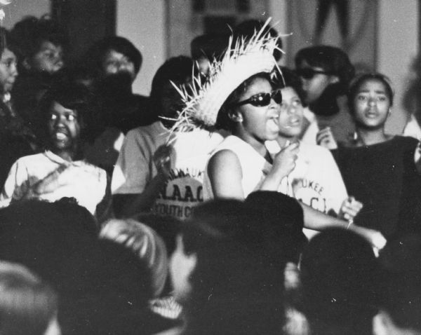 Group of people around a woman wearing sunglasses and a straw hat. A few of the people behind her are wearing NAACP shirts.