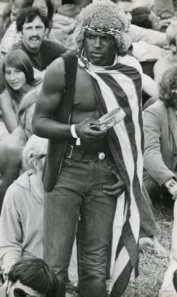 Elevated view of a man standing wearing a vest, furry hat and headband, American Flag cape and holding a tin of sardines in front of a crowd sitting at a rock festival at the "People's Fair."