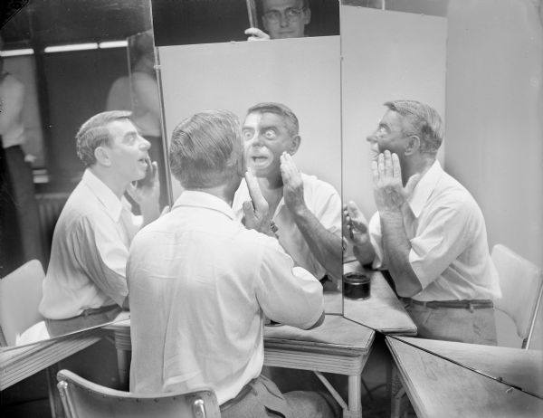 Eddie Cantor applying blackface makeup in front of a triple mirror. The mirror on the left reflects the left side of his face without makeup, the center mirror shows his face in half white, half black makeup, and the right shows the right side of his face in blackface makeup. A man is standing in the background behind the mirrors.