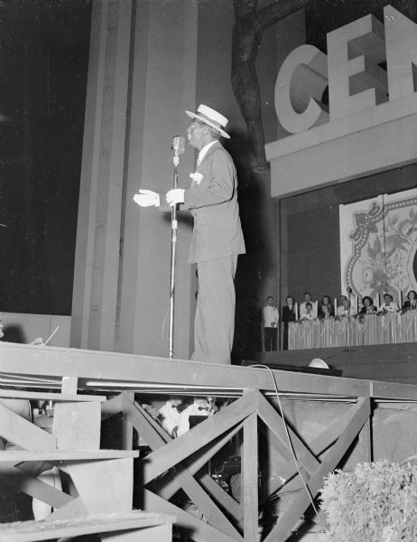 Eddie Cantor standing on stage wearing blackface. He is holding on to a microphone, and is facing towards the left. In the background audience members are sitting along a long table. The orchestra is sitting underneath the stage, and the conductor is standing with his head and hands and holding his baton just above the stage floor.