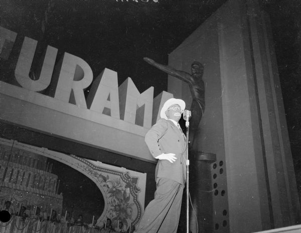 Eddie Cantor, in blackface, on stage at Centurama Evening Performance.  In the background men and women are sitting at a long table in front of a large image of a cake with candles.