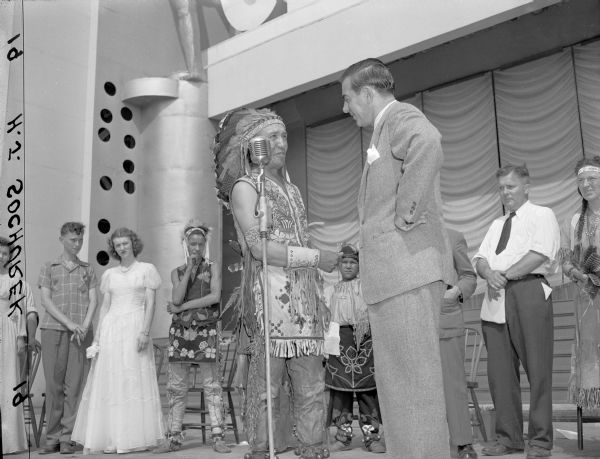 Eddie Cantor on stage at Centurama standing at a microphone with another man. People are standing in the background watching.