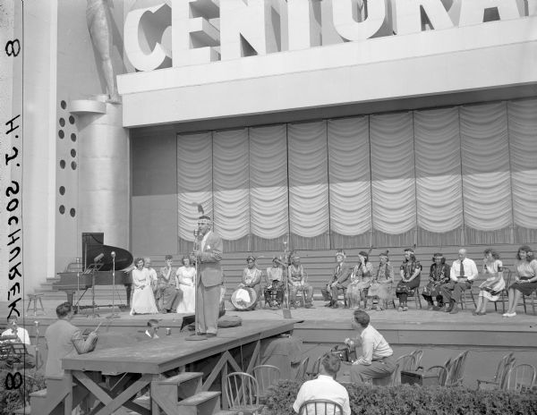 View of Eddie Cantor on stage standing in front of a microphone. There is a band below him on the left, and people are sitting in chairs on the stage behind him.