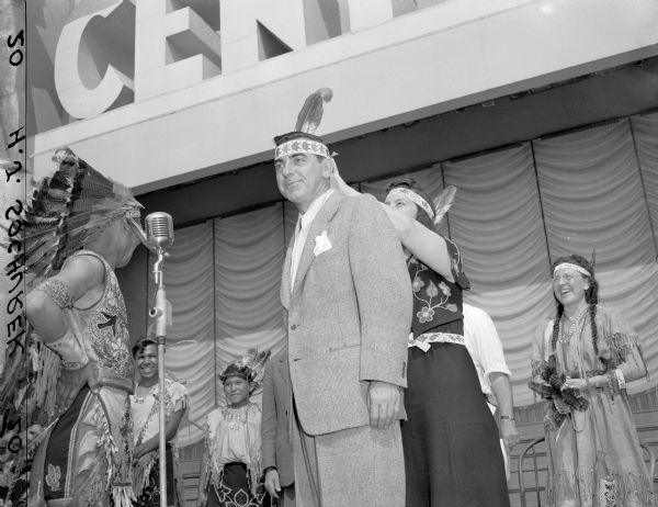 Man putting a feather and headband on Eddie Cantor, on stage at Centurama.