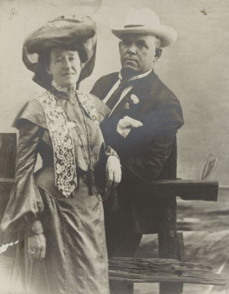 A souvenir photograph of Frank B. and Louise Morilla Mears Fargo, taken in Atlantic City. The couple is posing against a prop split rail fence in front of a backdrop which depicts a small sailboat on the ocean. Mrs. Mears is wearing a long sleeved dress with a capelet and short sash, and has a large hat with attached scarf. Mr. Mears is dressed in a suit and tie with a white hat and pocket handkerchief. The photograph is mounted on an album page and identified as "Atlantic City, July 1903, Mother Father."
